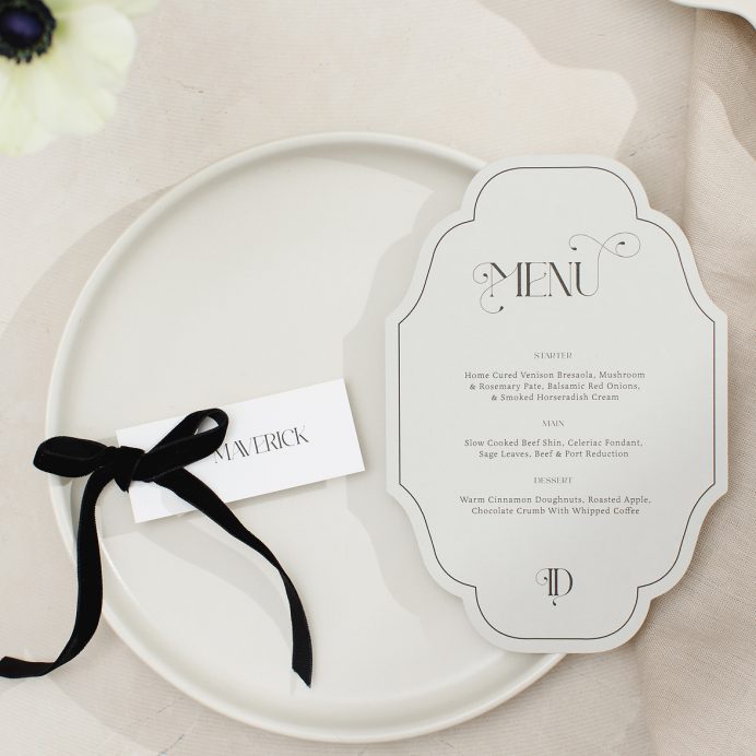 Vintage inspired cut to shape wedding menu in grey with ornate font. White guest name card with and black velvet bow.