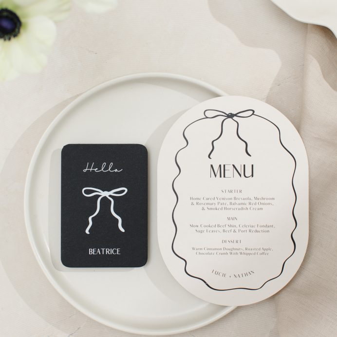 Double arch shape almond and black wedding menu with hand drawn squiggly line border and bow. Black wedding guest place card with white bow.